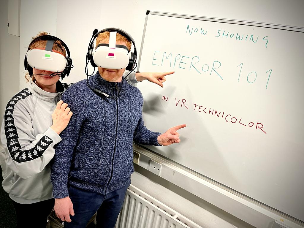 Performers Caitríona Ní Mhurchú and Karl Quinn stand pointing back at a white board. It says "Now Showing Emperor 101 in VR Technicolor"