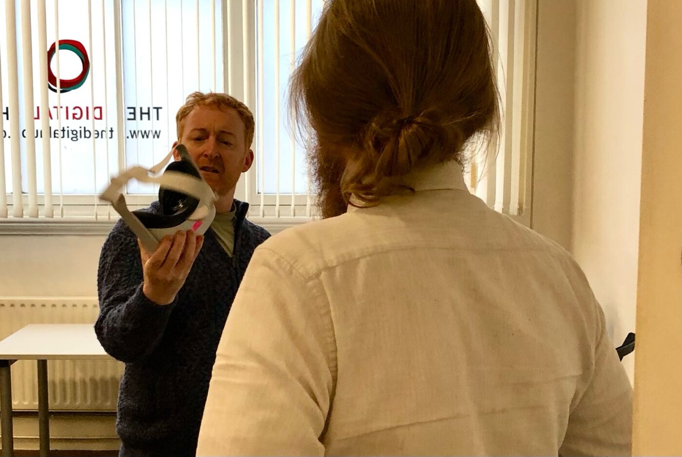 Performer Karl Quinn holds a VR Headset in his hand, looking perplexed. Fellow performer Caitríona Ní Mhurchú looks at him.