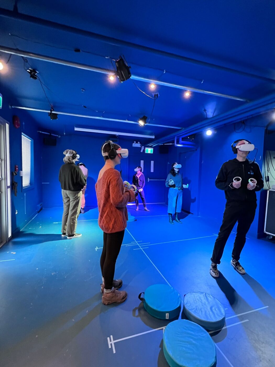 A crowd of 6 people stands in a blue room, each with a VR headset on and holding controllers, all looking in different directions.