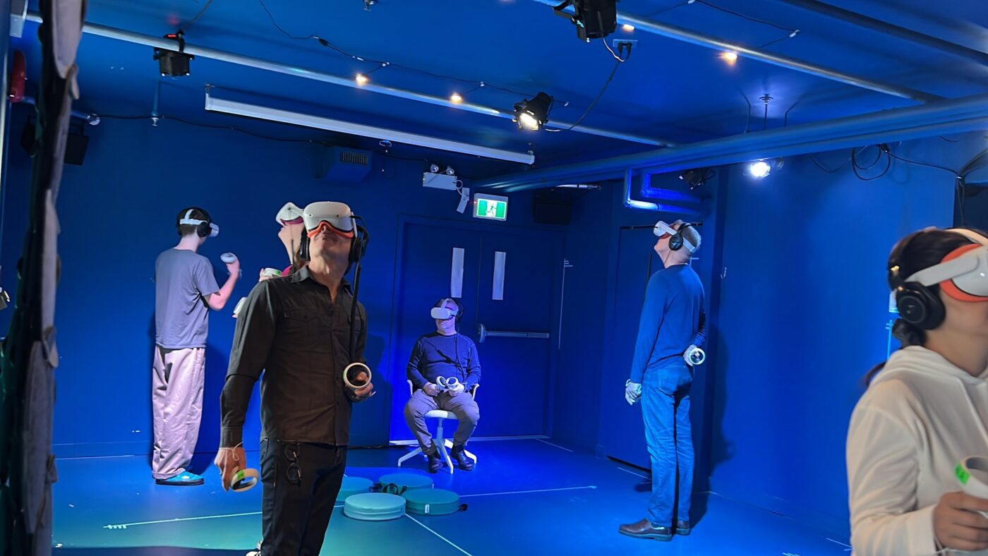 A group of people in a blue room wearing VR headsets look in a variety of directions, most of them looking up.