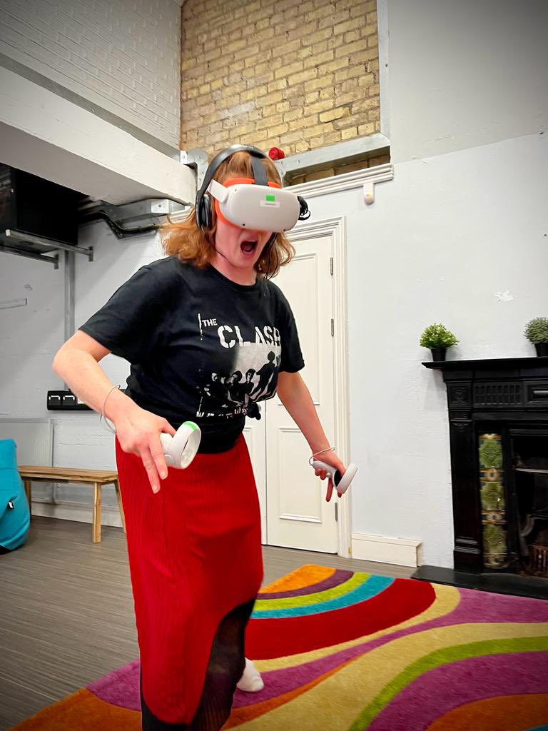 Caitríona Ní Mhurchú stands midperformance in a dramatic pose screaming at the audience. She wears her VR headset, controllers, comfy red pants, and a t-shirt from The Clash
