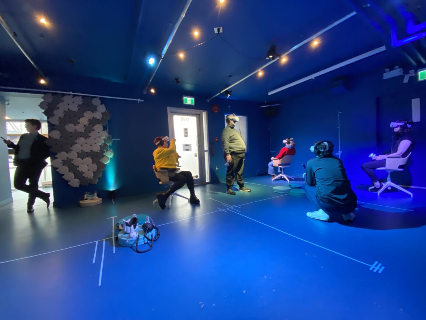 A group of 5 are in a blue room, some sitting, one standing, one kneeling, looking in different directions wearing VR headsets and controllers. Another figure leans against a door way, backlit.